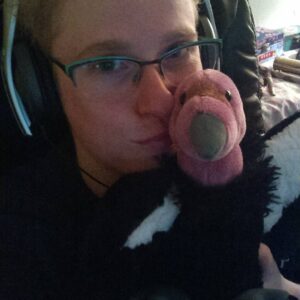 Photo of me in college. I am a white person with blue eyes and glasses, wearing large headphones and pensively cuddling with a vulture stuffie.