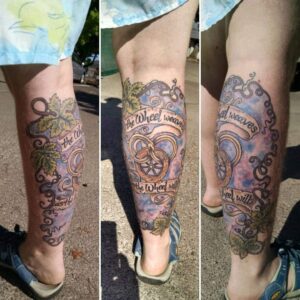 A tattoo on a calf that is of a snake weaving through itself and a wheel banned with text that reads "the Wheel weaves as the Wheel wills" and is surrounded by curling grape vines over a stormy sky.
